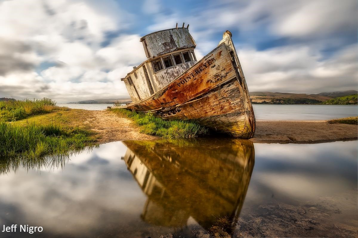 Photo of a shipwreck at low tide with its reflection appearing in the shallow water below.